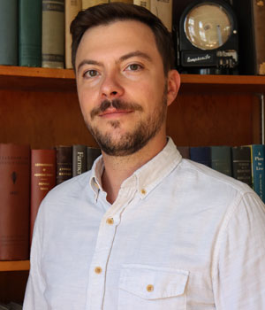 Headshot of Kyle Webert standing in a library with a vintage bookshelf and antique clock behind him.