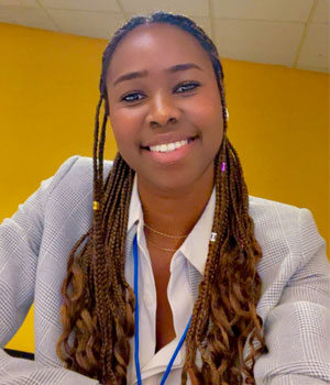 Headshot of Sylvia Onyeiwu female smiling and standing confidently with her arms crossed. She is wearing a white shirt and blazer.