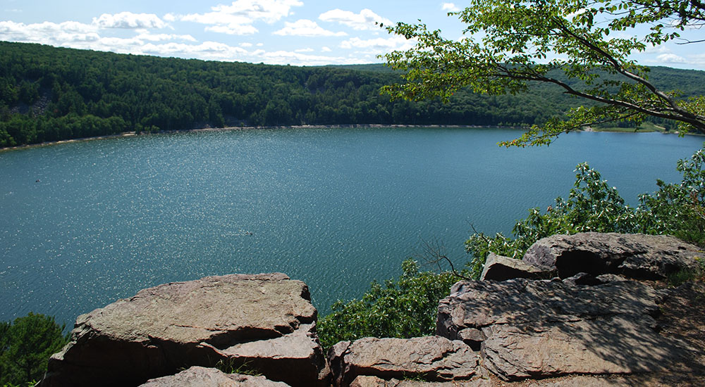 Photo of the East Bluff. There is a pond taking up the majority of the picture. The photographer is among a formation of rocks on the shore of the pond. Beyond the pond there is a heavy green forest