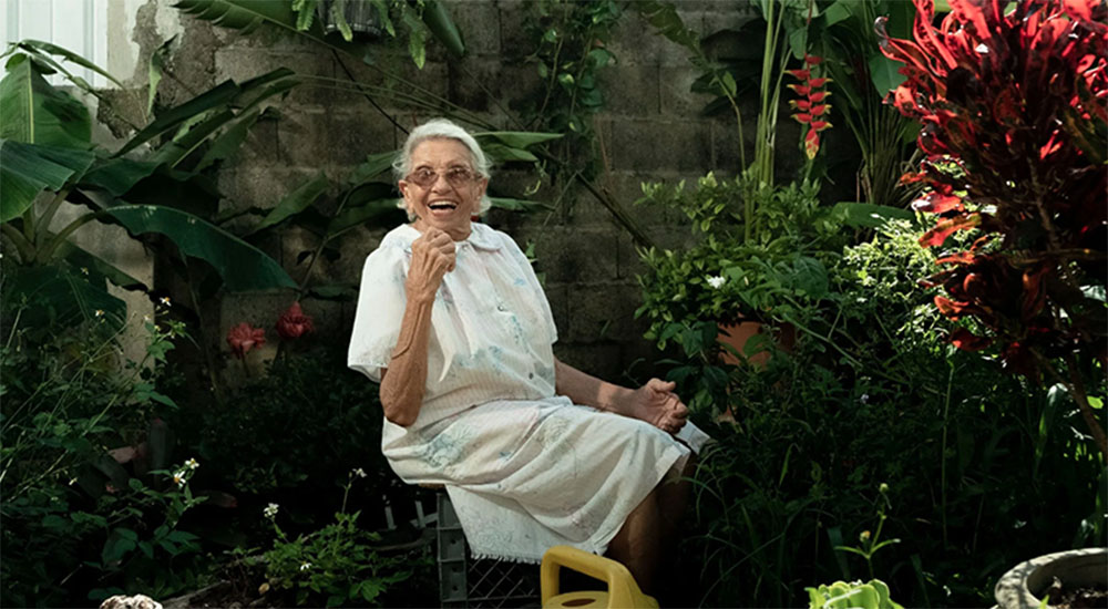 Photo from the movie "Gardenia Perfume". An old woman dressed in white sits in the middle of a grand garden full of greenery. She is smiling and holds a small flower in her hand.