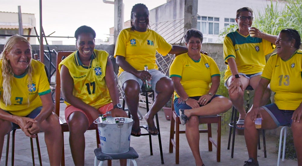 Photo from "The First Women". Six women stand in chairs in their soccer kits. They are all smiling at the camera, most of them have their arm on one another's shoulder to show their comradery