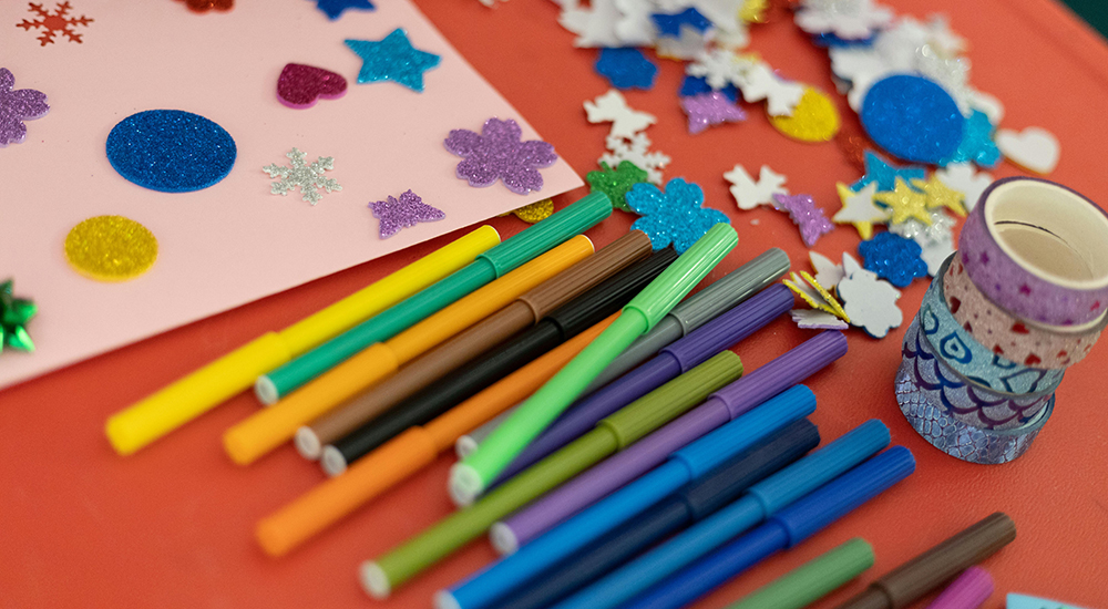 Table with a pile of skinny markers next to a piece of white construction paper with puffy stickers stuck on it. There are a pile of stickers beyond the markers.