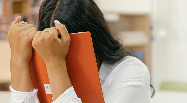 Photo of a woman holding a book with both hands and burying her face in the book. You cannot see her face, but her shoulders and the grip she has on the book hints that she is stressed