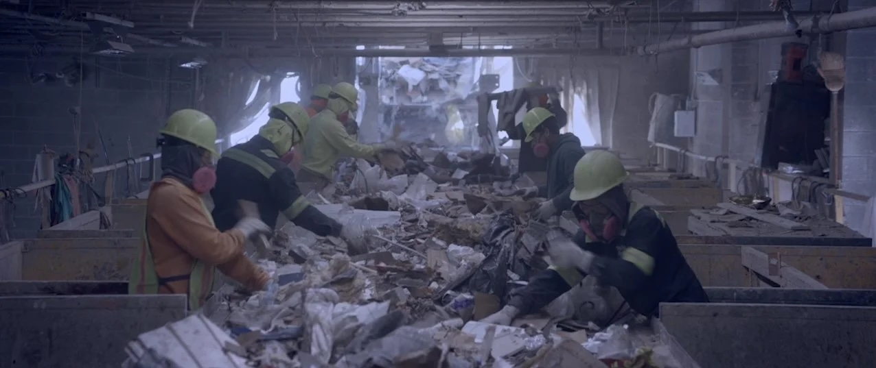 Photo from the documentary film "In Excess". Seven sanitation workers in hardhats are spread out along a table filled with trash as they sort it, seemingly from recyclable material and trash