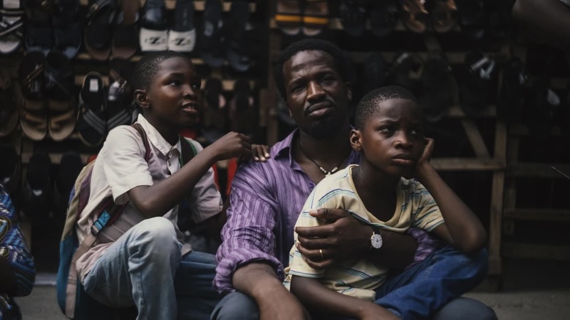 Photo from the film "Father's Shadow". A man sits with two young boys. One boy is sitting on the man's lap. Behind them are racks of shoes. The three people look tired, the boy in the man's lap his his head resting on his hand