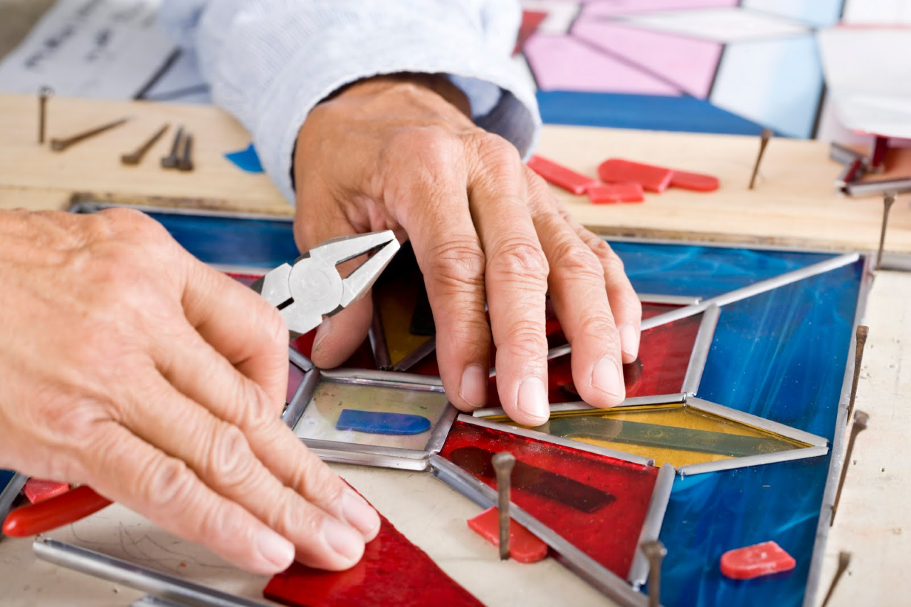 Close-up of a person's hands assembling a geometric stained glass panel using the lead came method. The hands use pliers to adjust a lead strip around vibrant red, blue, and yellow glass pieces, which are held in place by horseshoe nails on a wooden work surface.