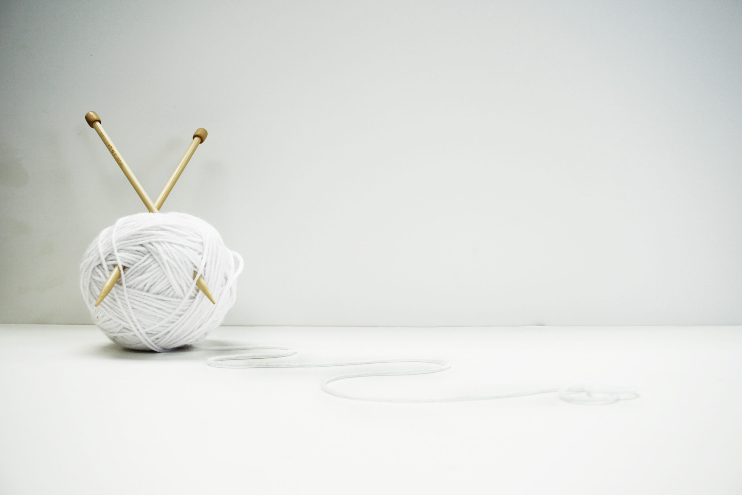 A ball of white yarn with two wooden knitting needles tucked through it, set against a clean, minimalist white background.