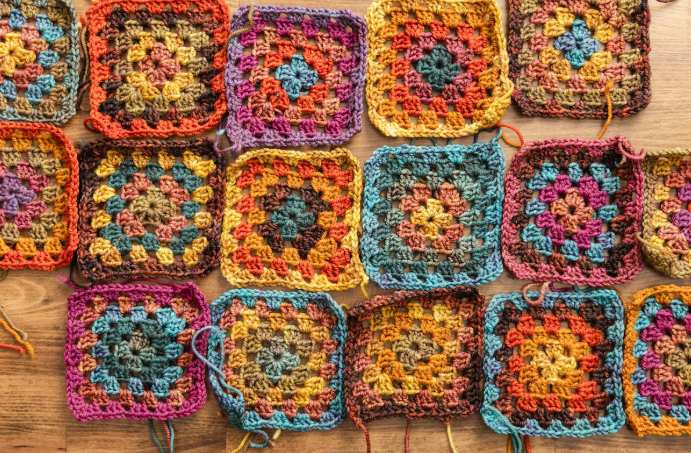 An overhead shot of several colorful, handmade crochet granny squares arranged in rows on a wooden surface, featuring various patterns of warm oranges, deep purples, and earthy blues.