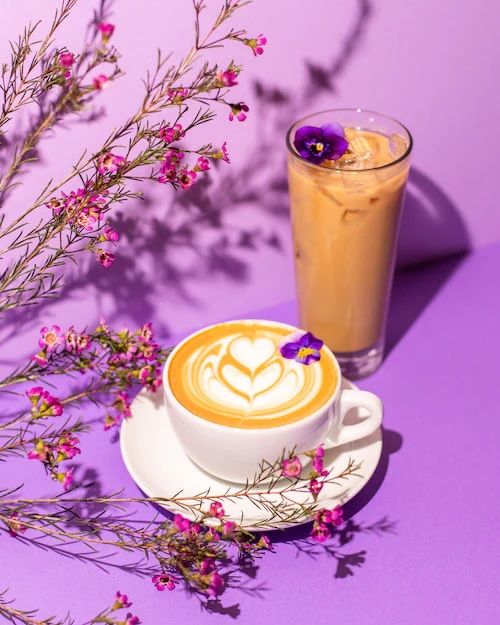 Photo of two coffee-based drinks and a bundle of lavender flowers. There is a flower petal in the latte mug. Latte art in the shape of a budding heart is in the cream of the latte. The taller drink is in a glass, it is tan in color with also a purple flower floating on top