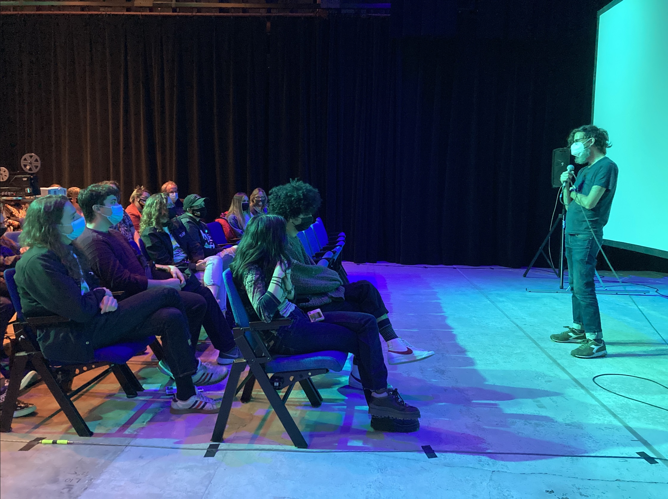 A presenter stands in front of a crowd of about 20 people sitting in folding chairs. A blue light is shining on the crowd and presenter, behind the presenter a white screen is set up.