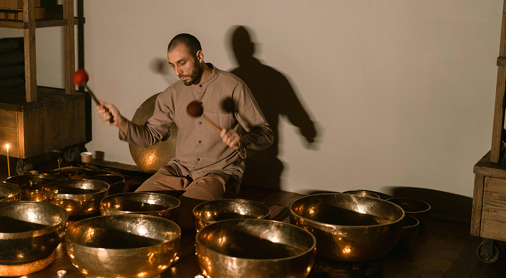 Photo of a man behind 8 large golden bowls he holds two mallets and his shadow is on the white background behind him. He is playing the bowls as an instrument