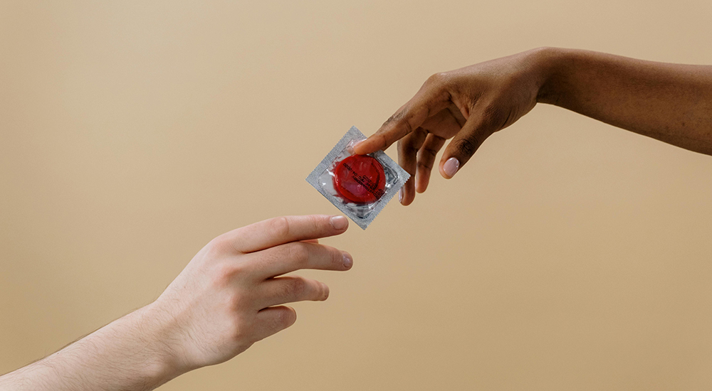 Photo of two hands, one coming from the bottom left of the photo and one from the top right of the photo. Their hands are meeting in the middle where they are appearing to exchange an unopened condom between each other
