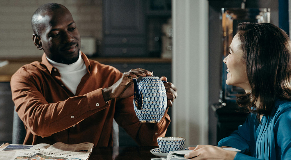 Photo of a man and woman sitting at a table sharing a pot of tea. The man is holding the tea kettle above the woman's cup, looking to fill it. They are both smiling, looking at each other