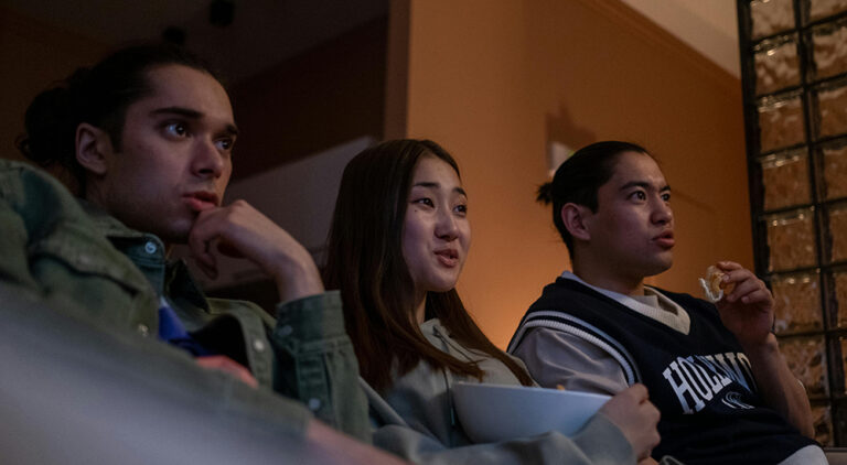 Photo of three people sharing a couch. The photo is taken from below, on an angle, toward their faces. All three seem to be enjoying whatever they are viewing together, they are attentive but not smiling, so it is not comedy. The person in the middle has a large bowl in their lap, so it appears they are also sharing a snack