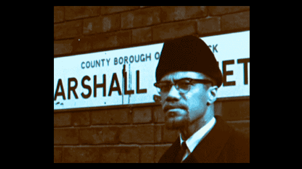 An image of Malcolm X standing in front of a street sign that reads Marshall Street in Smethwick, England. He is wearing a dark suit, glasses, and an Astrakhan hat, looking directly at the camera. The shot is a vintage, sepia-toned film still from the documentary Handsworth Songs.