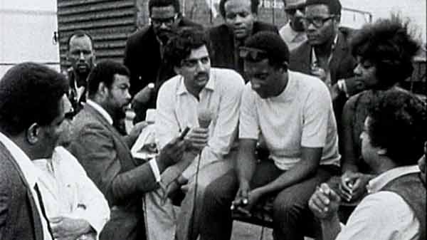 A grainy, black-and-white archival photograph from the film Who Needs a Heart, showing a group of Black activists and intellectuals gathered in an outdoor urban setting. In the center, two men are engaged in a serious conversation while others look on; a woman with an afro stands to the right. The scene captures the gritty, documentary-style aesthetic of the Black Audio Film Collective.