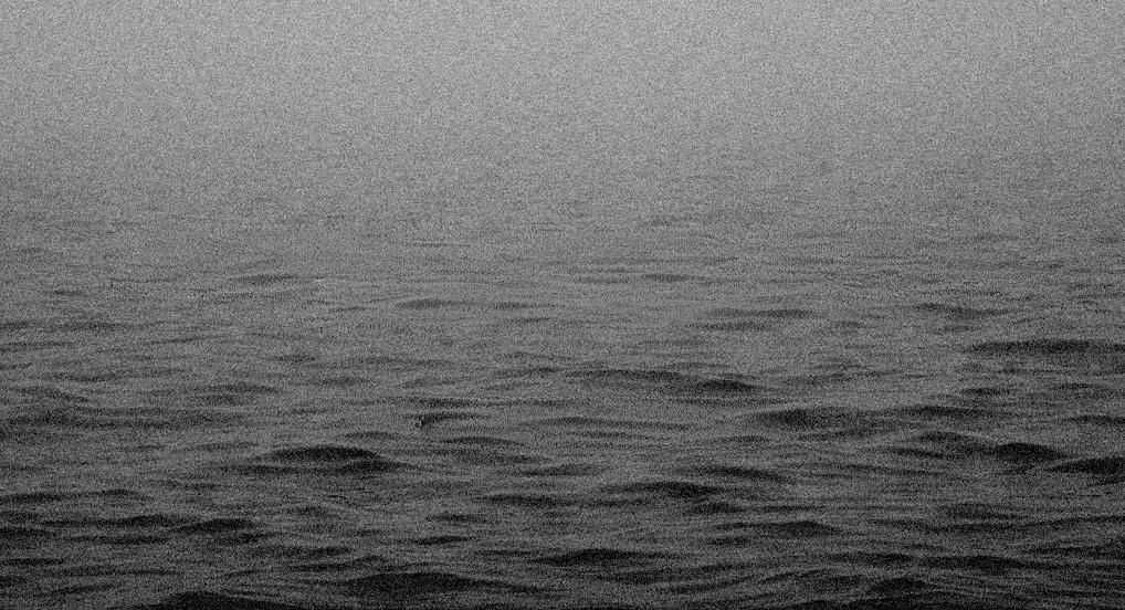A wide, panoramic shot of a dark, choppy body of water under a heavy, overcast mist. The horizon line is blurred by fog, creating a moody, monochromatic atmosphere.