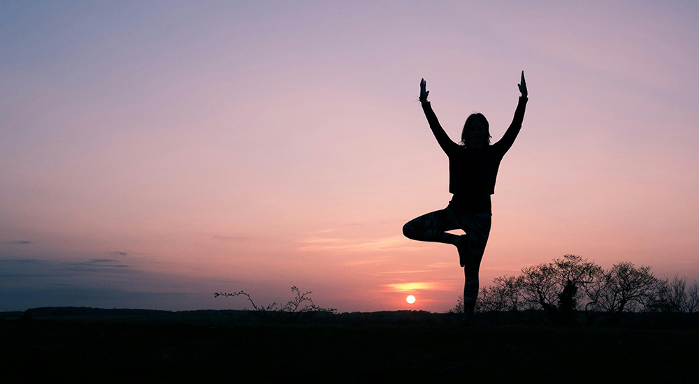 A silhouette of a person practicing Yoga (Tree Pose) against a vibrant sunset. The individual is balanced on one leg with their arms raised in a "V" shape, centered in the frame. The background features a soft gradient of purple and orange sky, with the dark outlines of trees and a flat horizon line below.