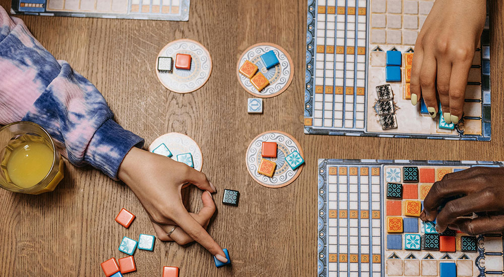 Bird's eye photo of a table with a board game being played on it. There are three pairs of hands hovering over individual game boards, and there are game pieces in the middle of the table. One hand is pointing towards a pile of game pieces