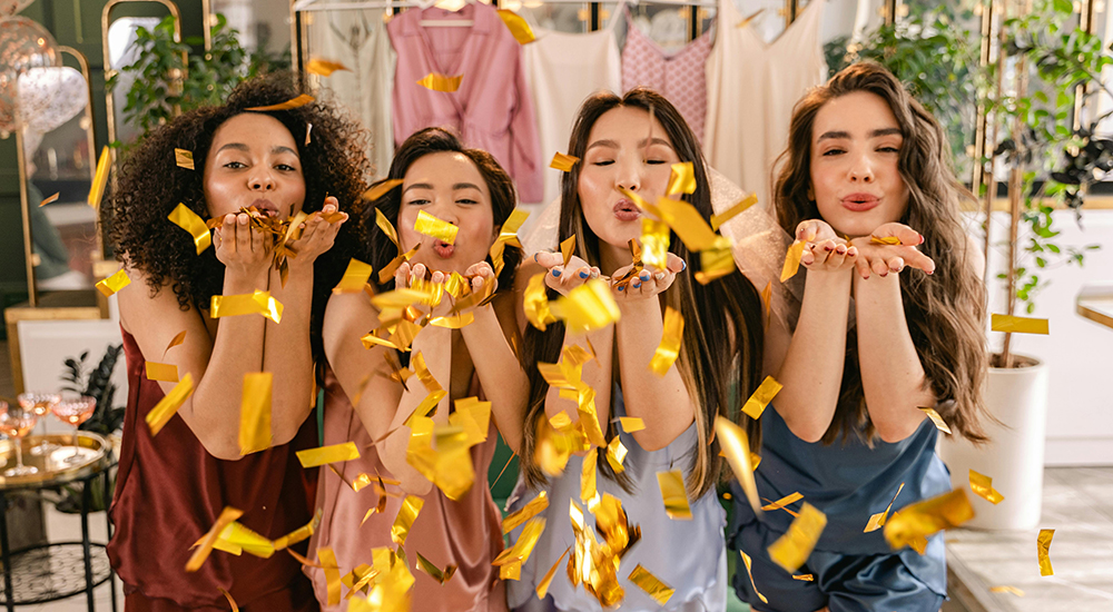 Four women in silk pajamas blowing gold confetti toward the camera in a brightly lit room.
