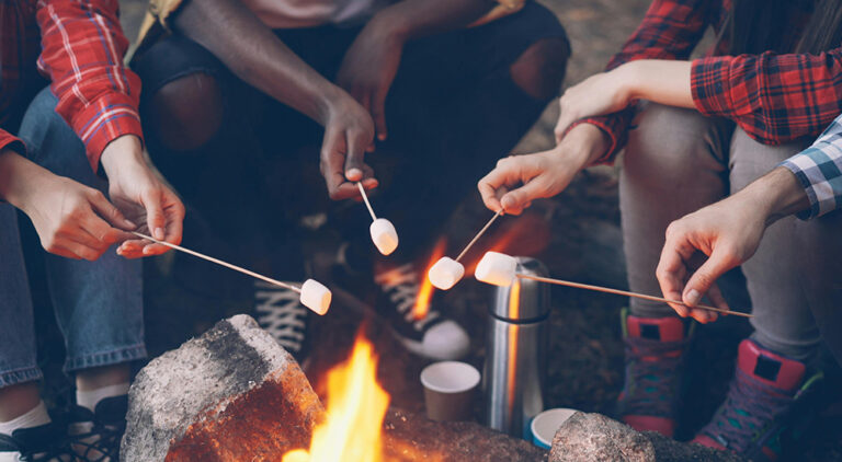 A group of people sitting around a glowing campfire at night, holding long skewers to toast marshmallows over the flames.