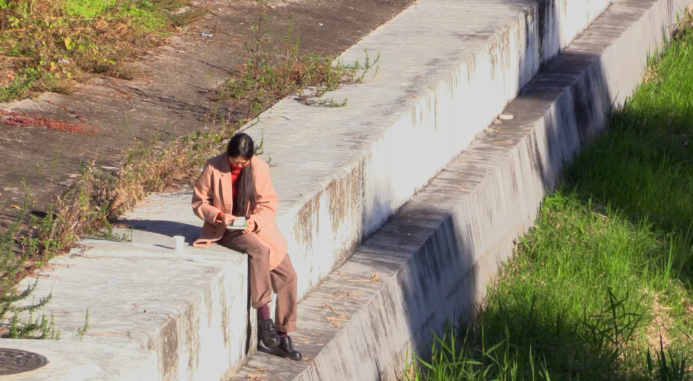 An overhead, long shot of a person sitting on the edge of a high, concrete embankment or retaining wall next to a grassy slope. The individual, dressed in a long beige coat and dark trousers, is looking down at a small object or device in their hands. The scene is captured in bright, natural sunlight, casting distinct shadows along the structured concrete lines.