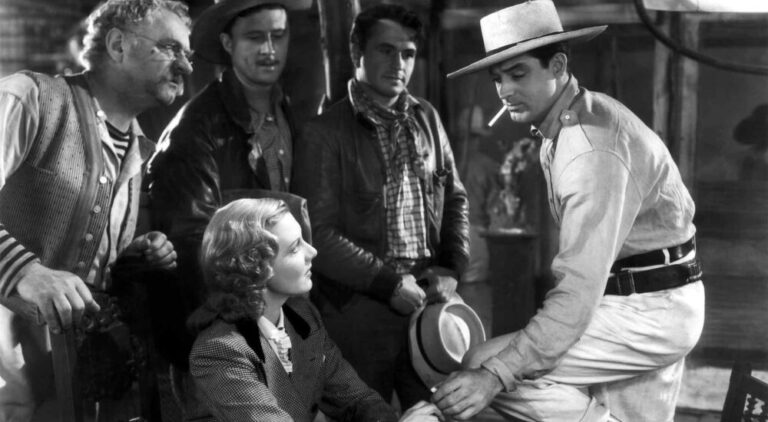 A black-and-white shot of a woman sitting down, surrounded by four men wearing hats and jackets, all looking toward a man on the right who is leaning forward and holding a hat.