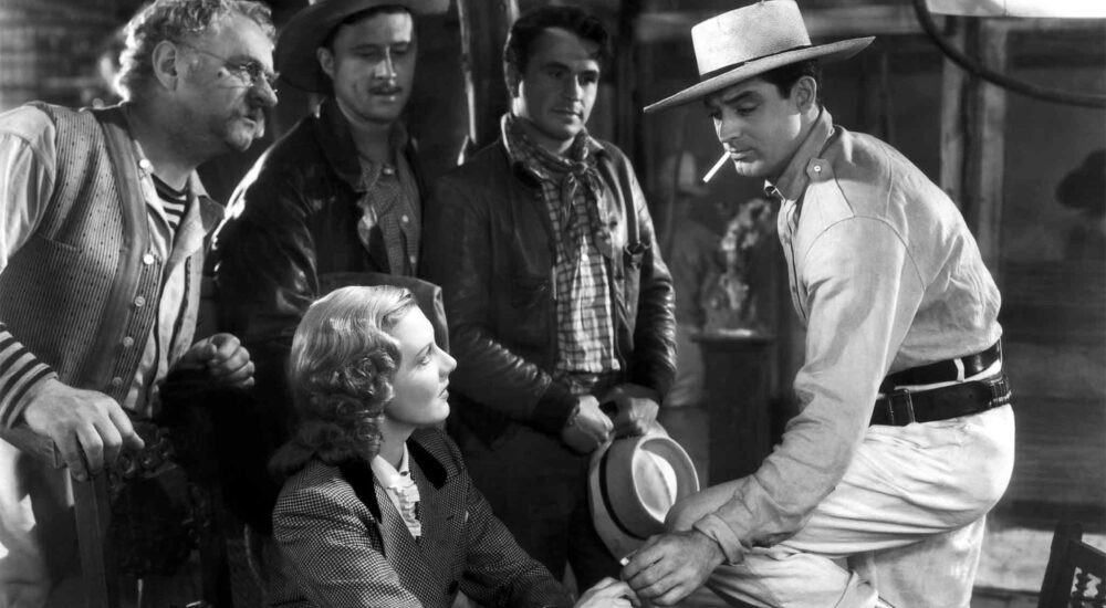 A black-and-white shot of a woman sitting down, surrounded by four men wearing hats and jackets, all looking toward a man on the right who is leaning forward and holding a hat.