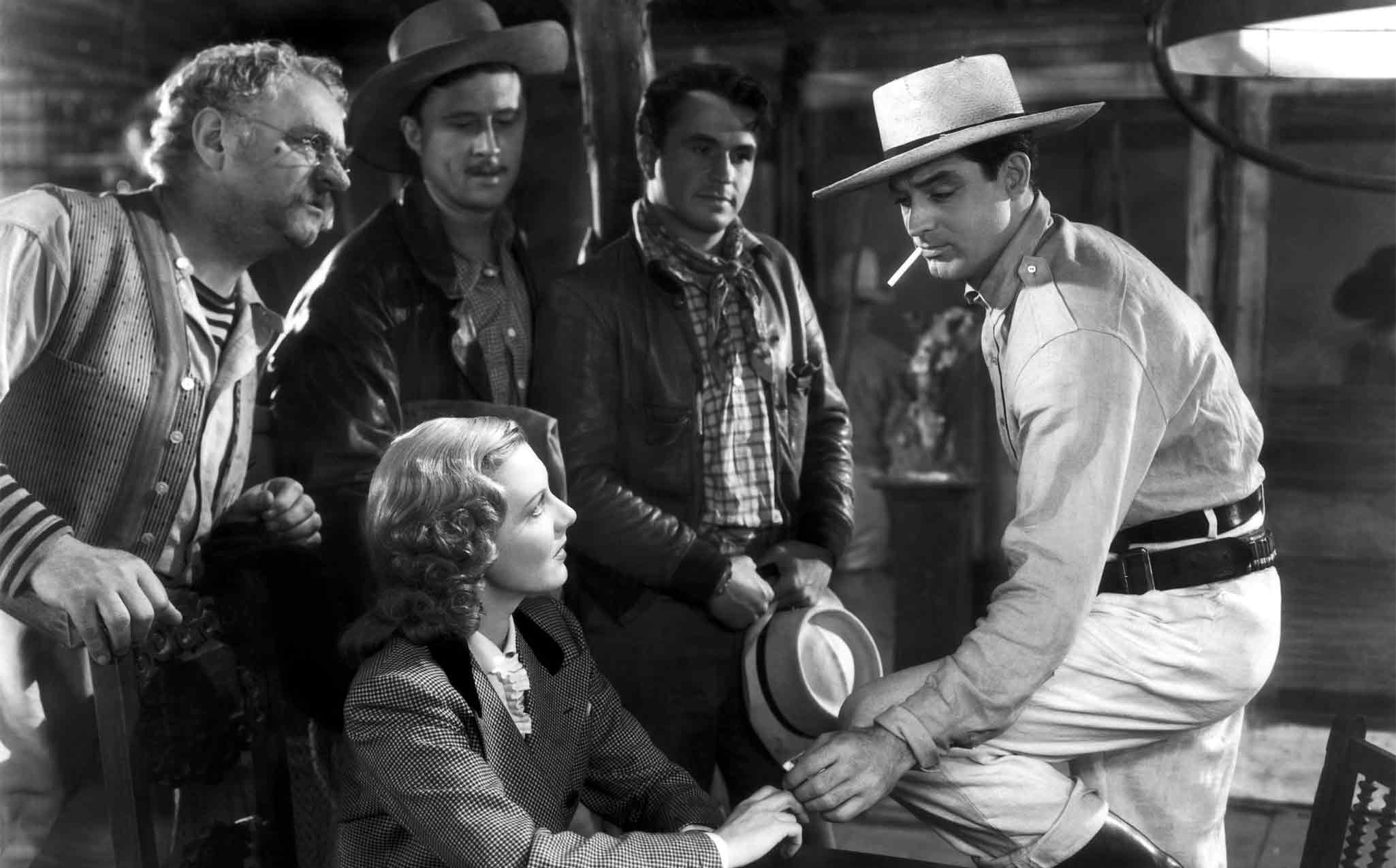 A black-and-white shot of a woman sitting down, surrounded by four men wearing hats and jackets, all looking toward a man on the right who is leaning forward and holding a hat.