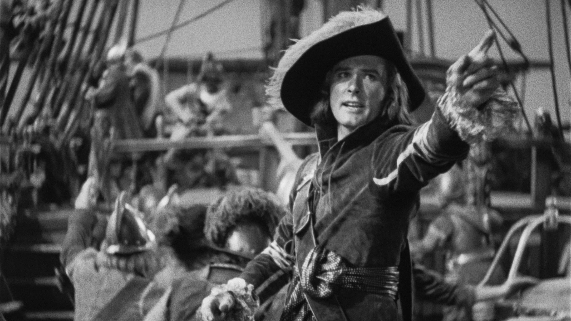 A black-and-white film still featuring Errol Flynn as Captain Blood on the deck of a ship, pointing dramatically forward with one hand while holding a sword or steering toward action. He wears a dark jacket, white ruffled cuffs, and a large, wide-brimmed pirate hat. In the background, rigging and other crew members are visible in soft focus.