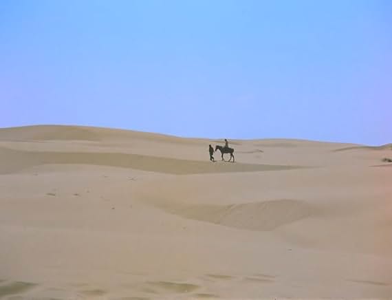 A wide, distant shot of two figures—one riding a horse and one walking beside it—traveling across vast, rolling desert sand dunes under a clear sky.
