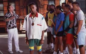 A medium shot from Spike Lee's film Do the Right Thing captures a group of people standing on a city street in front of a brick building. In the center foreground, Mookie stands looking forward, wearing a white short-sleeved button-up shirt with red and green collars, a large round medallion necklace, and green and yellow shorts. To his left, Sal stands in white pants and a black-and-white patterned shirt, gesturing with his hands while looking upward. To Mookie's right, a crowd of neighborhood residents, including Buggin Out and Radio Raheem, stand close together watching the interaction.