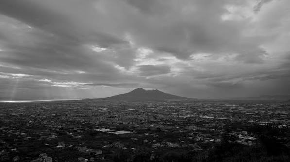 A wide-angle, black-and-white photograph capturing a vast expanse of land and a distant city spreading out toward a prominent, double-peaked mountain on the horizon. The sky is filled with a dramatic, heavy blanket of clouds, with distinct rays of sunlight breaking through on the far left, illuminating the edge of the landscape. The foreground features darker, elevated terrain looking down over the sprawling, densely patterned plains below.