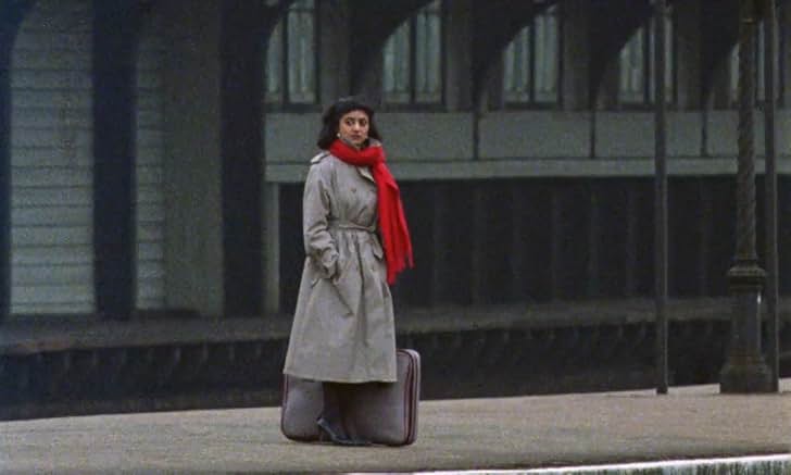 A wide, cinematic shot of a young woman standing alone on what appears to be a concrete train station platform or an outdoor pathway. She has short, dark hair and is wearing a long, light gray double-breasted trench coat belted at the waist, paired with a bright red scarf wrapped around her neck. A brown suitcase rests on the ground behind her. The background consists of a dark, long building with a row of arched windows or openings, creating a structured, geometric backdrop under a muted, overcast light.