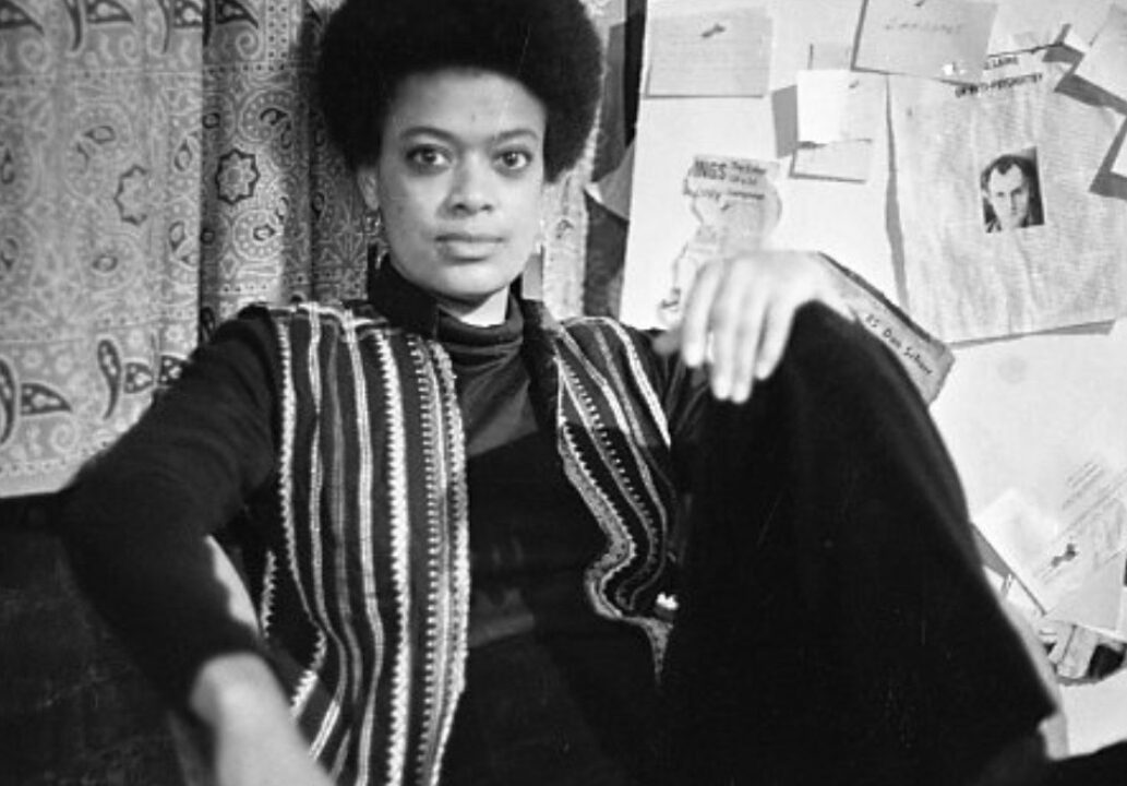 Black-and-white photograph of author and activist Toni Cade Bambara with an afro, wearing a patterned vest, sitting in front of a wall pinned with papers and clippings.