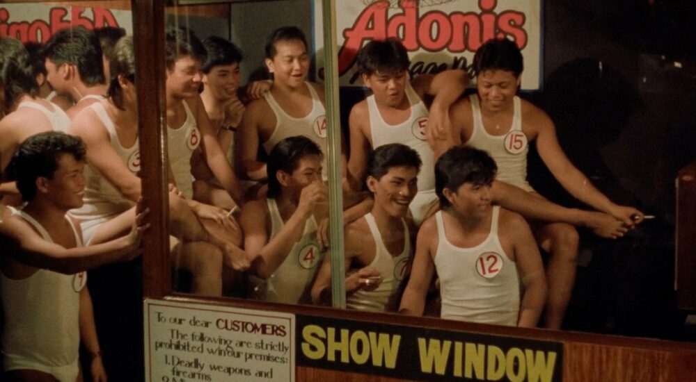 A group of young men wearing matching white tank tops and numbers pinned to their chests crowd behind a glass window labeled "SHOW WINDOW" under a glowing "Adonis" sign.