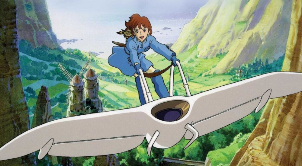 An anime still from Nausicaä of the Valley of the Wind showing Nausicaä, a young woman with short reddish-brown hair wearing a blue tunic and pants, flying her white, bird-like glider (the Mehve) through a lush green valley. She stands upright on the glider, holding onto its control bars, with a small, fox-squirrel creature perched on her shoulder. In the background, green cliffs and distant towers rise under a partly cloudy sky.