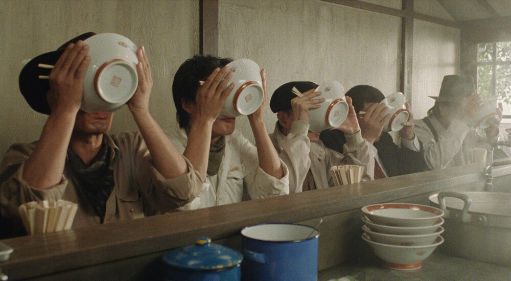 Five people sitting in a row at a restaurant counter simultaneously hold up large ramen bowls, completely covering their faces as they drink the broth.