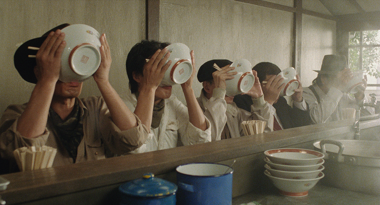 Five people sitting in a row at a restaurant counter simultaneously hold up large ramen bowls, completely covering their faces as they drink the broth.