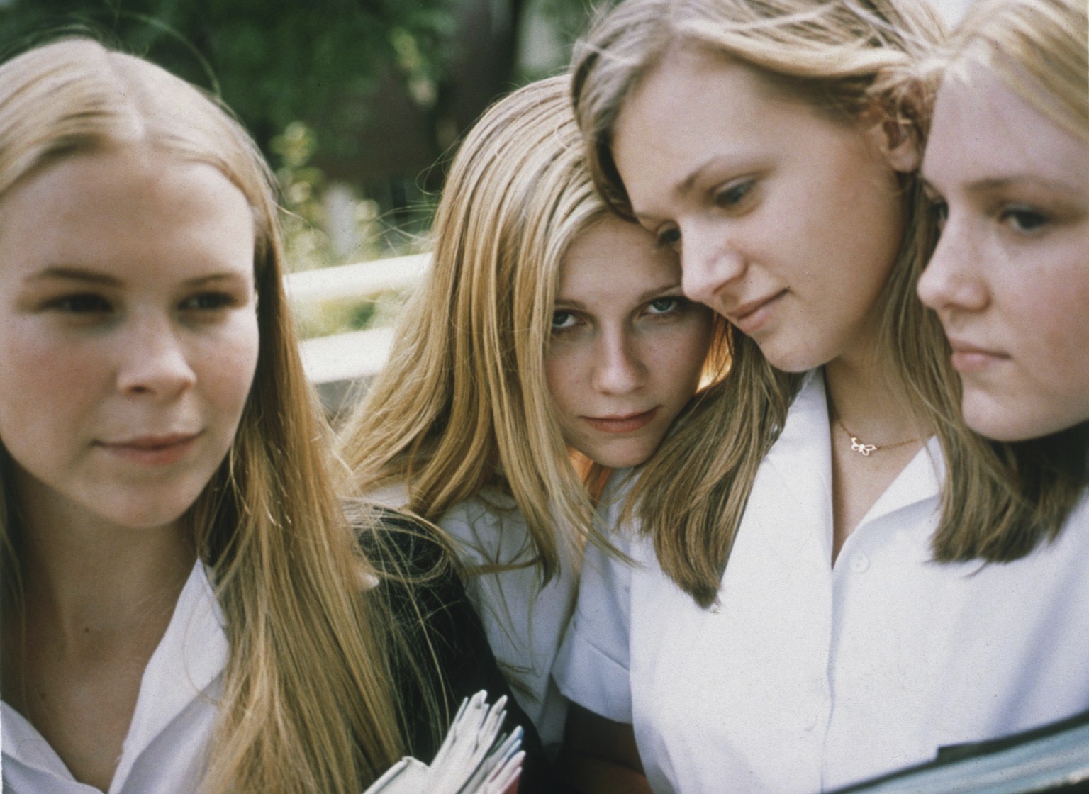 A close-up shot from Sofia Coppola's film The Virgin Suicides captures four young women with long blonde hair clustered tightly together. They are dressed in matching white, short-sleeved collared shirts, looking in different directions with subtle, somber expressions. One woman in the lower-left corner holds open a book or binder, which is partially visible at the bottom of the frame.
