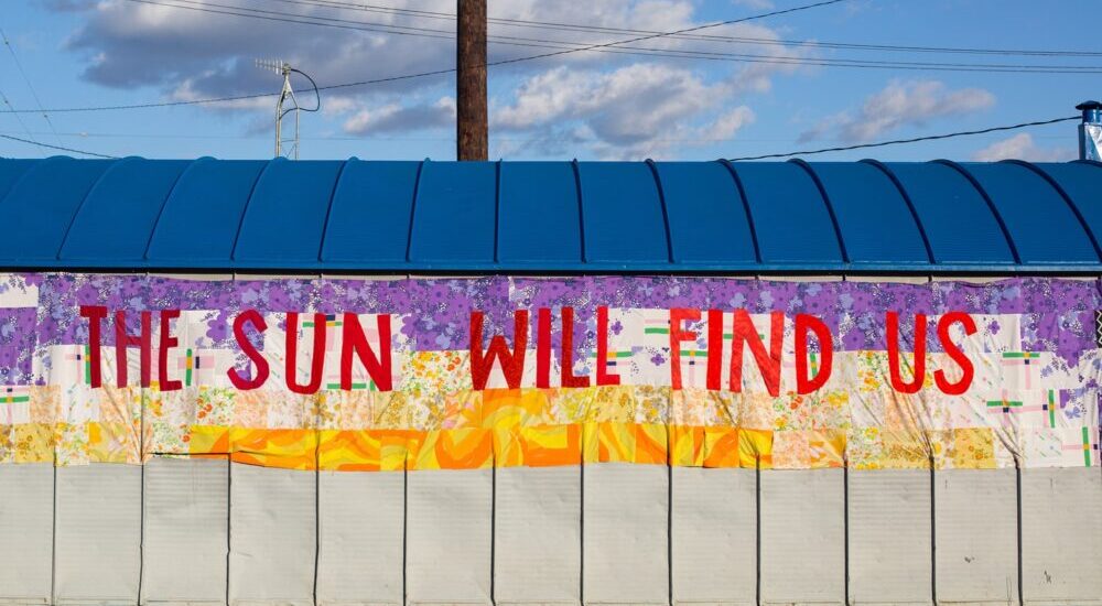 A low-angle shot of a wall painted with the phrase "THE SUN WILL FIND US" in red lettering below a blue-roofed structure.