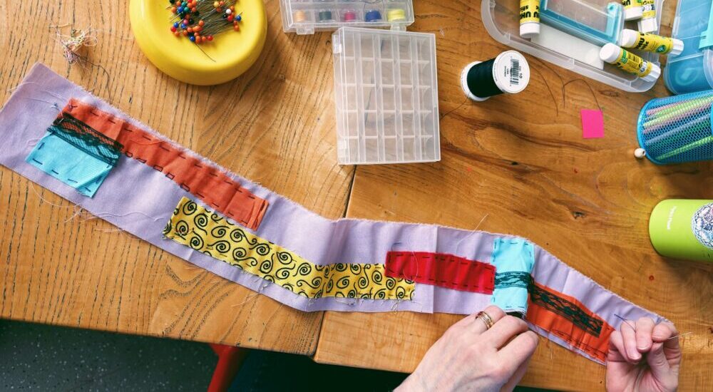 An overhead view of a wooden workbench with sewing supplies and a hands-on fabric project. Hands at the bottom hold a needle and thread, working on stitching colorful patterned fabric patches onto a long strip of light purple cloth. Surrounding the workspace are a yellow pincushion with colorful pins, clear organizer boxes with thread spools, spools of black thread, and glue sticks.