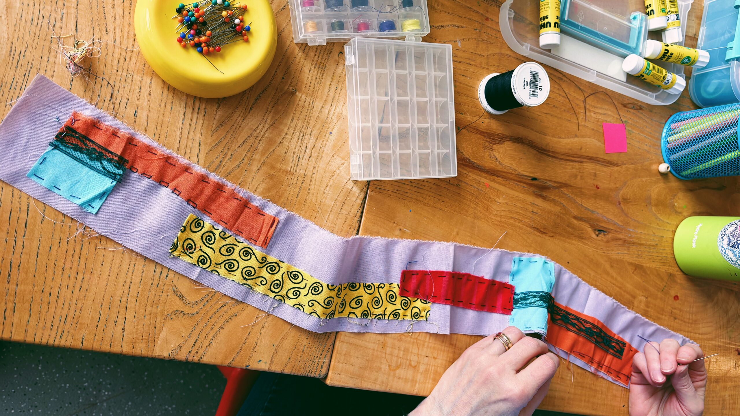An overhead view of a wooden workbench with sewing supplies and a hands-on fabric project. Hands at the bottom hold a needle and thread, working on stitching colorful patterned fabric patches onto a long strip of light purple cloth. Surrounding the workspace are a yellow pincushion with colorful pins, clear organizer boxes with thread spools, spools of black thread, and glue sticks.