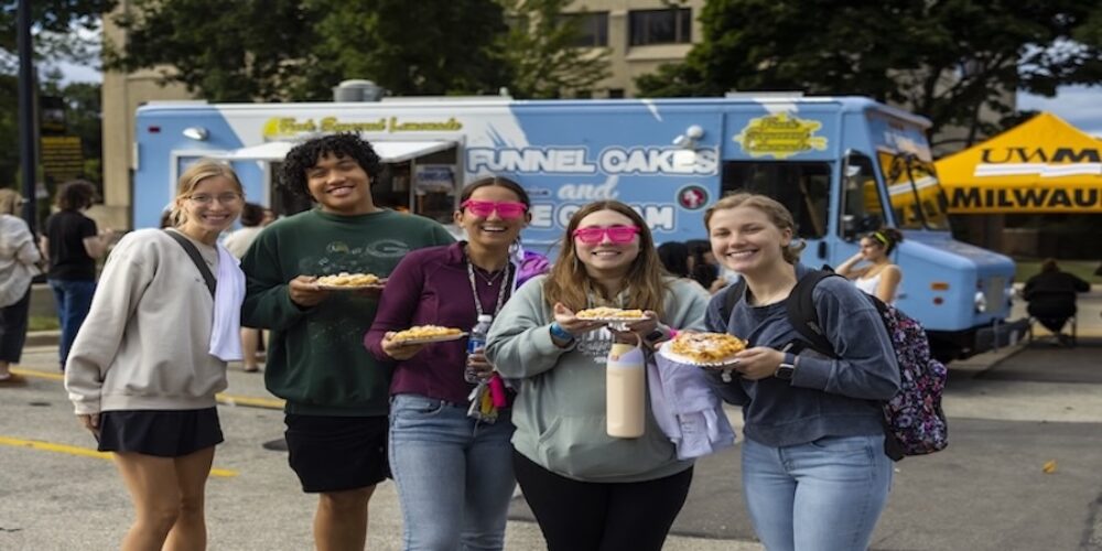 A group of five smiling young adults standing together outside on a sunny day, holding plates of food in front of a blue "Funnel Cake" food truck.