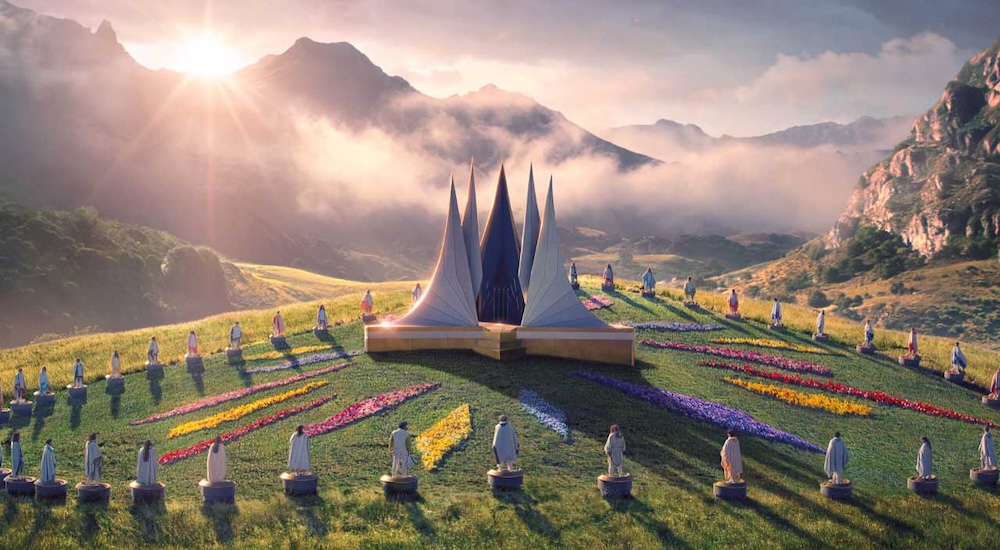 A digital panoramic cinematic shot depicting a sunlit, grassy valley surrounded by misty mountains. In the center sits an arena-like layout with an architectural structure featuring tall, dark, pointed spires arranged in a circle. Radiating outward from this central structure are long, manicured rows of vibrant, multi-colored flowers. A large group of small figures stands in a wide semicircle facing the structure on the green field.