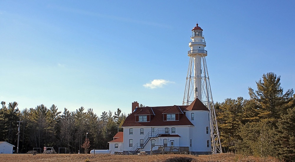 A skeletal steel light tower painted white stands tall next to a two-story white keeper's quarters with a red roof, situated in a clearing surrounded by evergreen pine trees against a clear blue sky.