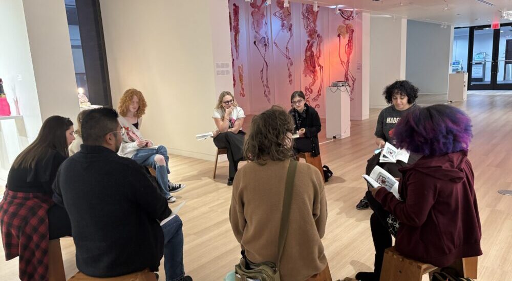 A group of people sits in a circle on wooden stools in a modern art gallery space, participating in a group discussion or reading while holding booklets, with pink artwork hanging on the wall in the background.
