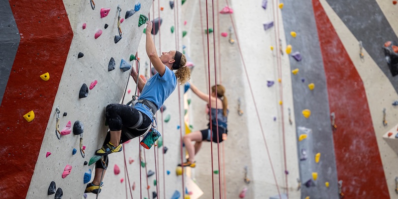 Two people scale a tall indoor rock climbing wall fitted with colorful handholds and vertical safety ropes.