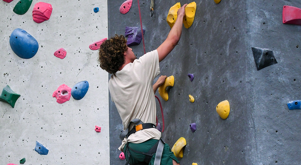 A young man with curly brown hair scaling an indoor climbing wall, viewed from behind and slightly below. He wears a beige t-shirt, green shorts, and a climbing harness attached to a red belay rope. His hands and feet are positioned on bright yellow climbing holds along a textured dark gray wall section, situated next to a light gray wall section featuring pink, blue, green, and purple holds.