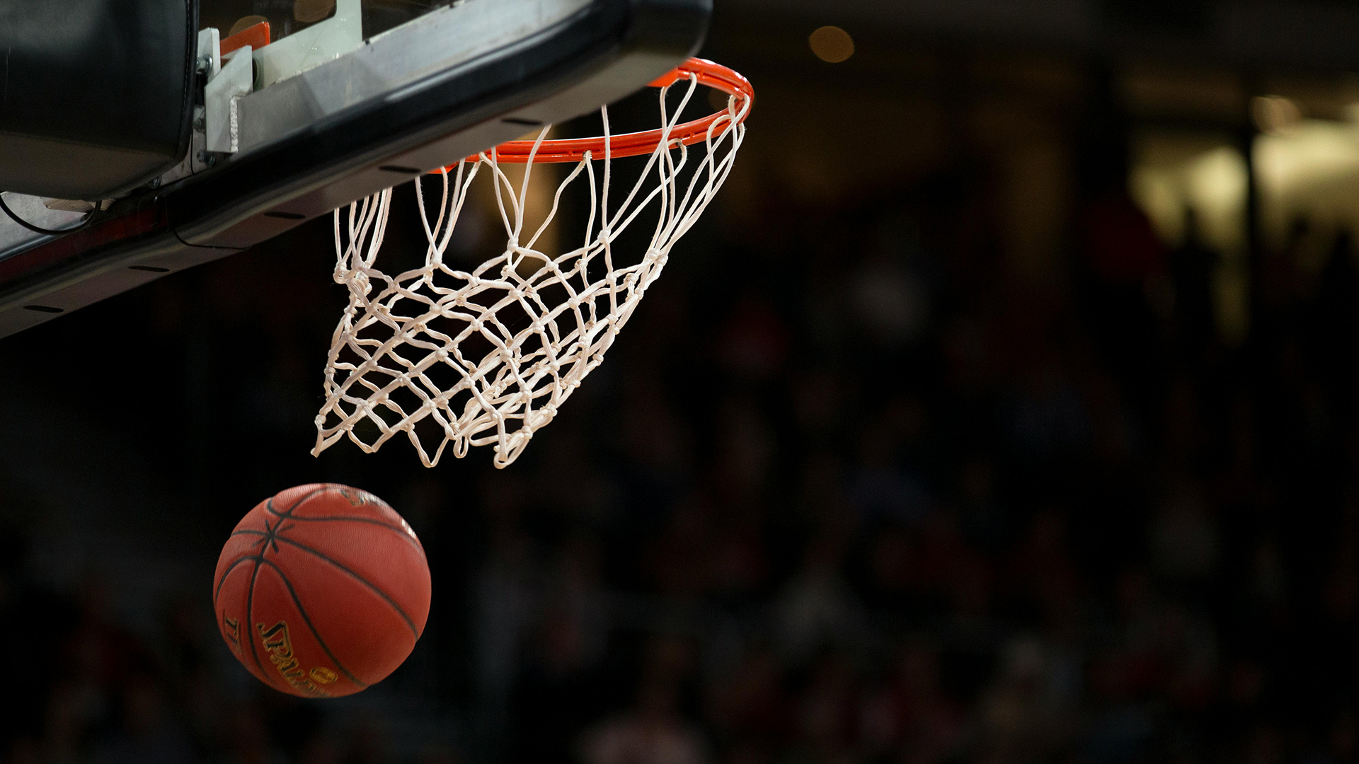 A low-angle, close-up shot of a basketball falling through an orange hoop and white net, set against a dark, blurred arena background.